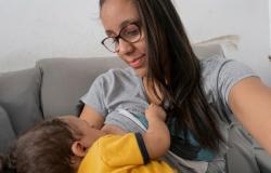 Mulher de cabelos longos e escuros, de óculos e camiseta cinza, amamenta um bebê de camiseta amarela. Os dois estão sentados em um sofá. A mulher sorri enquanto olha para o bebê, que mama e segura sua camiseta.