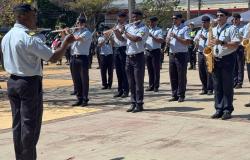 Banda da Guarda Civil Municipal de Belo Horizonte se apresenta em praça pública sob regência de maestro, que está de costas na imagem, tendo em frente vários músicos tocando instrumentos de sopro. Todos estão uniformizados.