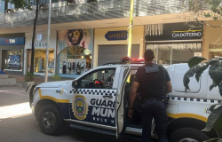 Viatura da Guarda Municipal de frente para três lojas, na rua. Agende da Guarda, uniformizado, parado ao lado da viatura, que está com a porta traseira aberta.