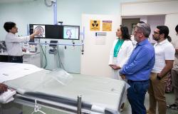 Imagem mostra uma maca em primeiro plano e atrás dela um homem apontando para duas telas de um aparelho do Serviço de Trombectomia Mecânica. O secretário de Saúde, de camisa azul, obserba, ao lado de uma mulher de camisa verde e branca. Há um homem de camiseta branca.