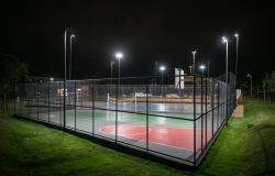 Quadra de esporte pintada de verde, vermelho e azul, cercada por tela, sem nenhuma pessoa, num ambiente noturno que ressalta a iluminação com postes.