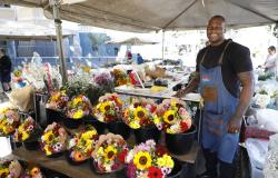 Barraca de flores em uma feira, com três andares de buquês com flores amarelas, vermelhas, rosas e brancas, ao lado do feirante Rogério da Silva, que está à direita, sorri e usa avental azul e blusa preta. 