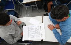 Imagem de dois alunos sentados frente à frente em carteiras escolares e olhando para um papel com atividade