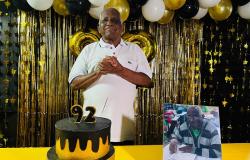 Imagem mostra o senhor José Maria Borges com as mãos postas em frente ao seu bolo de aniversário, que tem uma vela de 92 anos e é dourado e preto, mesmas cores dos bações e da cortina que estão no fundo. Há também uma foto dele em sala de aula.