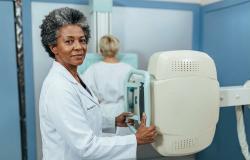 Foto de uma sala de exames de saúde. Em primeiro plano, uma profissional de saúde negra, de cabelos grisalhos e curtos, veste um jaleco branco e segura o equipamento de mamografia. À direita, aparece o aparelho de mamografia, de cor branca, com uma estrutura vertical e uma área de apoio para posicionamento da paciente. Ao fundo, uma mulher, de cabelos claros e usando roupa branca, está de costas diante do equipamento. 