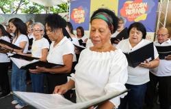 Apresentação do coral do projeto Saúde Encanto no Centro Cultural Urucuia. Em primeiro plano, uma mulher negra idosa veste blusa branca de ombro a ombro e faixa colorida no cabelo, segurando uma pasta preta de partitura aberta e cantando com os olhos direcionados para a folha. Ao lado e atrás dela, outros integrantes do coral, vestindo roupas brancas, acompanham a música com suas pastas. Ao fundo, há um painel colorido com a inscrição "11º Descontorno Cultural".