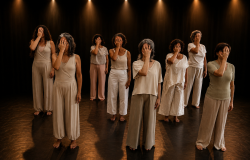 Oito mulheres estão em pé sobre um palco escuro, sob focos de luz amarelada, organizadas em duas fileiras. Todas vestem roupas em tons neutros e claros (bege, off-white e cinza-claro) e cobrem o próprio rosto com uma das mãos, espalmada sobre os olhos. O ambiente transmite uma atmosfera dramática e performática. 