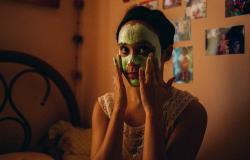 Jovem mulher com o rosto coberto por uma máscara facial verde aplicando o produto com as mãos, em cena do filme Medusa. Ela está em um quarto aconchegante com iluminação alaranjada e fotos na parede ao fundo.