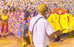  Fotografia colorida em plano médio de uma apresentação cultural festiva. Em primeiro plano, centralizado e de costas para a câmera, um homem vestindo camisa xadrez clara, chapéu de palha e calça jeans segura uma baqueta com a mão direita e carrega um tambor a tiracolo. 