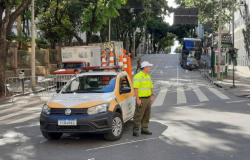 Agente de trânsito usando uniforme da BHTrans, nas cores verde e amarelo, com boné branco, ao lado de veículo usado em operação de trânsito, parado em via de BH