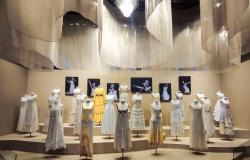 "Fotografia da exposição 'Clara Nunes - Eu Sou a Tal Mineira'. Em uma sala com iluminação suave e teto decorado com tecidos ondulados, vários manequins exibem figurinos originais da cantora. A maioria são vestidos longos em tons brancos e cremes, com detalhes em rendas e franjas, além de um vestido amarelo ao centro-esquerda. Ao fundo, quadros em preto e branco mostram fotos de Clara Nunes no palco."