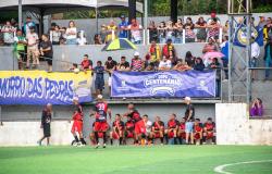 Fotografia de uma partida da Copa Centenário de Futebol Amador. Em primeiro plano, jogadores de uniforme preto e vermelho estão próximos ao banco de reservas, ao lado do campo de grama sintética.
