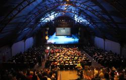  Fotografia panorâmica do interior de um teatro, registrada do fundo da plateia em direção ao palco. O espaço está lotado, com centenas de pessoas sentadas em poltronas organizadas em fileiras e algumas circulando pelos corredores. O público é formado, em grande parte, por pessoas idosas. O teatro possui teto alto e arqueado, com estrutura metálica aparente, iluminado em tons de azul. Ao fundo, o palco está iluminado por luzes azuis e apresenta um efeito de fumaça. Acima dele, uma tela exibe a inscrição “Sa