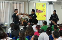 Na imagem há três guardas municipais. Dois fazem uma apresentação, enquanto uma policial feminina observa. A plateia é formada por alunos de uma escola municipal.