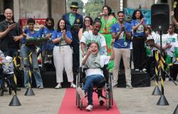 Fotografia de um evento ao ar livre. No centro, uma criança com deficiência está em uma cadeira de rodas, sobre um tapete vermelho, acompanhada por uma mulher de camiseta azul que conduz a cadeira. A criança segura um adereço verde. Ao redor, várias pessoas acompanham o momento, algumas sorrindo, aplaudindo e gesticulando. Ao fundo, há um palco com participantes, microfones e cartazes coloridos.