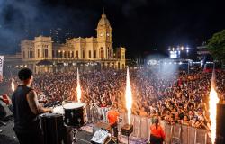Um músico toca tambores no palco diante de uma grande multidão animada, com braços erguidos, na Praça da Estação, em Belo Horizonte. Jatos de fogo iluminam o ambiente, enquanto, ao fundo, destaca-se o prédio histórico do Museu de Artes e Ofícios, com sua torre de relógio.