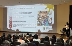 Imagem mostra um auditório com pessoas de costas e uma mulher no palco apresentando um power point com informações sobre alimentação nutricional.