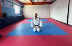 Em uma sala de treinamento de artes marciais, o professor Igor Oliveira posa, ajoelhado, em um tatame azul e vermelho. Igor usa quimono branco. Ao fundo e ao lado, há paredes brancas e azuis.m.
