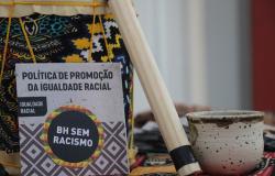 A imagem mostra uma composição de objetos sobre um tecido colorido com estampas geométricas de inspiração africana. À esquerda, há um tambor revestido com tecido estampado e cordas amarelas. À frente, está apoiado um material impresso com os dizeres “Política de Promoção da Igualdade Racial” e “BH Sem Racismo”, acompanhado de grafismos geométricos em diferentes cores. Ao centro, um objeto cilíndrico de madeira clara está posicionado na diagonal. 