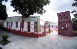 Fachada do Centro Cultural Salgado Filho. A imagem destaca o totem de identificação na cor vinho com o nome do centro esculpido em preto, e a grade metálica vermelha que cerca a área externa. Ao fundo, o prédio moderno com paredes verdes claras e janelas com detalhes de vidros coloridos (vermelho, amarelo, azul). 