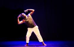 Homem negro dança descalço no centro de um palco escuro, sob iluminação amarelada e chão azul. Ele veste regata preta e calça branca. Inclina o tronco para a direita com as pernas abertas, joelhos dobrados e as mãos espalmadas ao redor da cabeça, em expressão concentrada.