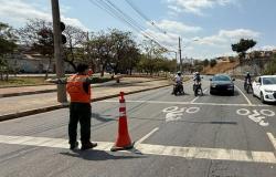 Imagem mostra um agente da Defesa Civil com colete laranja orientando o trânsito. Na foto há três motoqueiros parados e dois carros. Há árvores do lado esquerdo.