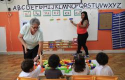 Duas professoras, uma de blusa vermelha e outra de blusa verde-clara, auxiliam quatro crianças em uma atividade com blocos coloridos sobre uma mesa, em uma sala de aula da Escola Municipal Marconi.