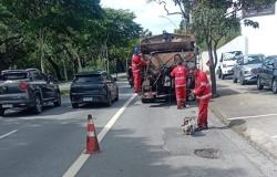 A iamgem mostra três homens com uniformes vermelhos em obras de tapa-buracos. Um deles está em um caminhão tanque e outros dois no asfalto. Ao lado do caminhão há dois carros supostamente em transito e do lado direito outros três supostamente estacionados.