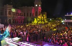Imagem noturna capturada do alto de um palco na Praça da Estação, durante um evento da Virada Cultural. Em primeiro plano, veem-se as treliças metálicas do palco e grades de proteção que separam a área de apresentação de uma multidão densa e entusiasmada, iluminada por feixes vibrantes de luzes rosa, amarela e verde. Ao fundo, destaca-se o prédio histórico da antiga Estação Ferroviária.