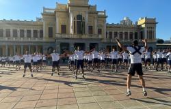 Fotografia ao ar livre, em dia ensolarado, mostrando um grande grupo de pessoas realizando exercícios físicos na praça da Estação.Todos estão vestindo uniforme esportivo composto por camisetas brancas de manga curta e calções escuros. De costas para a câmera, um instrutor salta com braços e pernas abertos conduzindo o exercício de polichinelo, enquanto os participantes estão posicionados em fileiras organizadas ao fundo acompanhando o mesmo movimento.