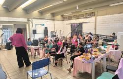 Imagem de uma sala com palestra para um grupo de mulheres. À esquerda, a palestrante veste blazer rosa e calça preta, vista de costas. No centro, cerca de 15 a 20 mulheres estão sentadas em cadeiras assistindo à palestra. À direita, há uma mesa com toalha rosa servida com comidas e bebidas.