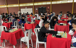 Estudantes participam de competição de xadrez em um espaço amplo, com mesas cobertas por toalhas vermelhas e tabuleiros organizados para as partidas. Os jovens estão sentados em cadeiras, concentrados nos jogos, enquanto outros participantes conversam e acompanham a atividade ao fundo.