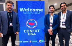 Três homens vestindo ternos azuis, sendo um deles com camisa preta e os outros dois com camisas sociais brancas, posando ao lado de um cartaz com fundo azul com os seguintes dizeres: Welcome Confunt USA. 