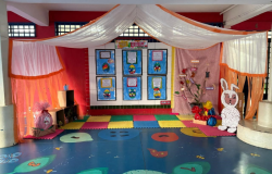 #paratodosverem: a foto mostra um cenário de contação de histórias com elementos lúdicos e pedagógicos. O ambiente está organizado com tapetes coloridos no chão, painéis com produções infantis nas paredes e uma decoração com tecidos leves. À direita, há uma árvore feita com galhos secos e nomes colados, além de um coelho decorativo.