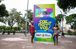 Totem colorido do Carnaval de Belo Horizonte, nas cores amarelo, verde, roxo e azul, com a marca “Carnaval de BH” e contador eletrônico que marca a contagem regressiva de 100 dias para a folia. Em frente ao totem duas mulheres, caracterizadas com roupa carnavalesca em posição de dança.