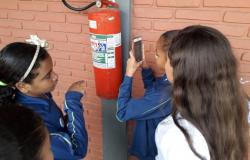 Quatro estudantes fiscalizam extintor de incêndio em escola municipal, durante o dia. 