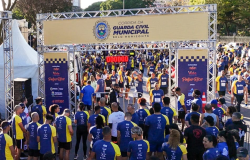 Multidão em frente a uma estrutura de metal com banner da Guarda Municipal se preparando para iniciar a corrida 