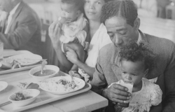Em foto P&B de acervo do Museu Histórico Abílio Barreto, homem em frente à mesa alimenta criança que está sentada em seu colo.