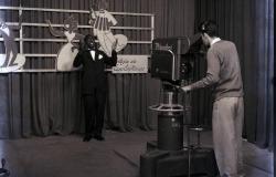 Foto em preto e branco feita em um estúdio de televisão. Um homem está em frente a uma câmera enquanto outro manuseia o equipamento