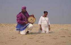 Homem negro ajoelhado toca tambor ao lado de mulher negra com roupa branca ajoelhada, na areia, durante o dia. 
