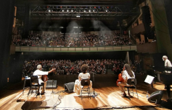 TReês artistas negros se apresentam no palco; ao fundo, uma platéia lotada assiste. 