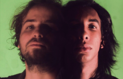 Dupla de rock com dois homens lado a lado, com os lados de dentro da imagem sombreados, dando a impressão de uma parcial fusão de rostos. 