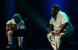O cantor BNegão ao lado de um homem com violão, ambos sentados, fazendo apresentação musical. 