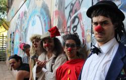 Seis pessoas, com roupas circenses, posam para foto encostados em muro grafitado, durante o dia