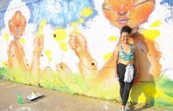 Mulher posa à frente de muro grafitado, durante o dia. 