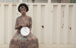 Foto do trabalho de Dayane tropicaos, com mulher negra de vestido, sentada em frente à portão, com um relógio na mão. 