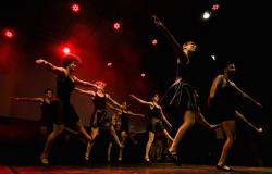 Sete bailarinos, quatro mulheres com saia e camiseta pretas, e os três homens com com short e camiseta pretos, dançam, de maneira coordenada, em palco teatral com luz vermelha..