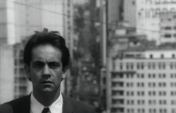 Imagem em P&B do ator Paulo José, muito jovem, de terno, com prédios ao fundo. Cena do filme A Vida Provisória.