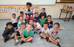 #paratodosverem: a foto mostra crianças em uma sala de aula. Com exceção de uma menina, a turma está sentada no chão. A aluna, ao centro, senta-se em uma cadeira e segura o boneco Tobias, que tem pele negra e usa blusa azul clara, shorts, calçados e uma boina azul escura. Ao fundo, há um quadro branco e cartazes com palavras acompanhadas de uma ilustração.