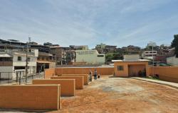 A foto mostra uma área pública aberta, com piso de terra e trechos pavimentados, cercada por muros baixos pintados de laranja. Ao fundo, há uma área residencial densamente ocupada, com casas de diferentes tamanhos e estilos, muitas construídas em alvenaria aparente, distribuídas por uma encosta. No centro da imagem, algumas pessoas circulam pelo espaço, enquanto, à direita, há uma pequena edificação de cor alaranjada, com portas e janelas protegidas por grades.
